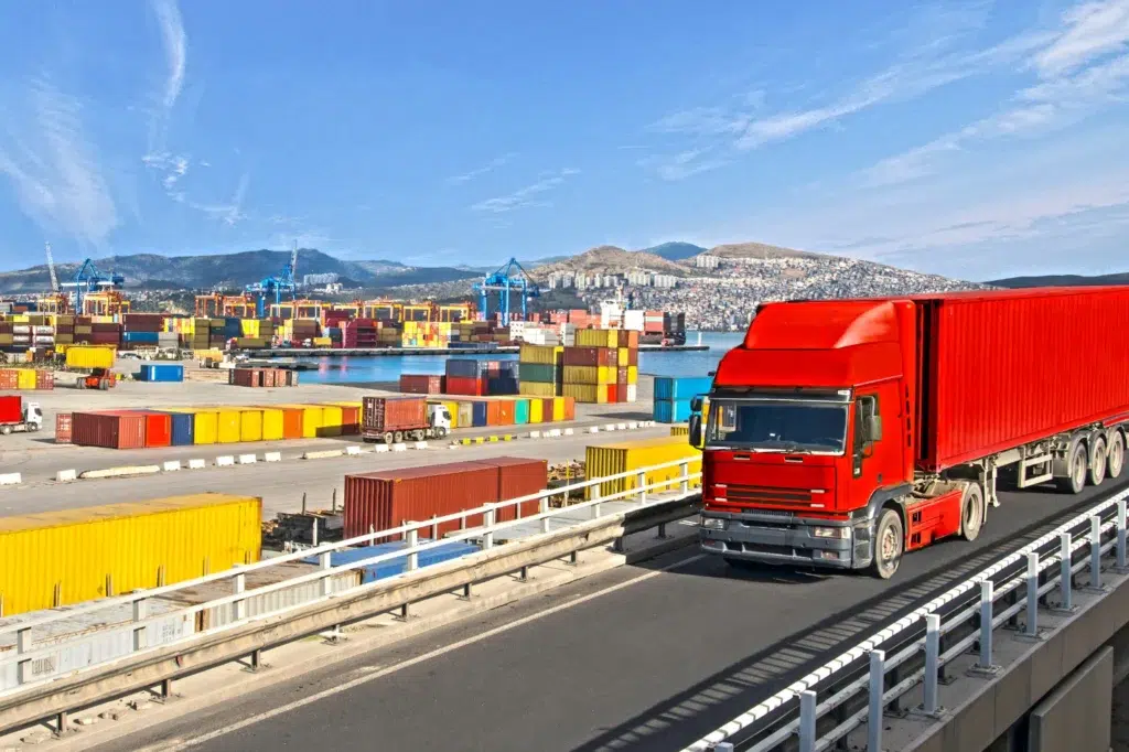 Red truck carrying a shipping container from a port crowded with cranes and colorful containers in central Europe